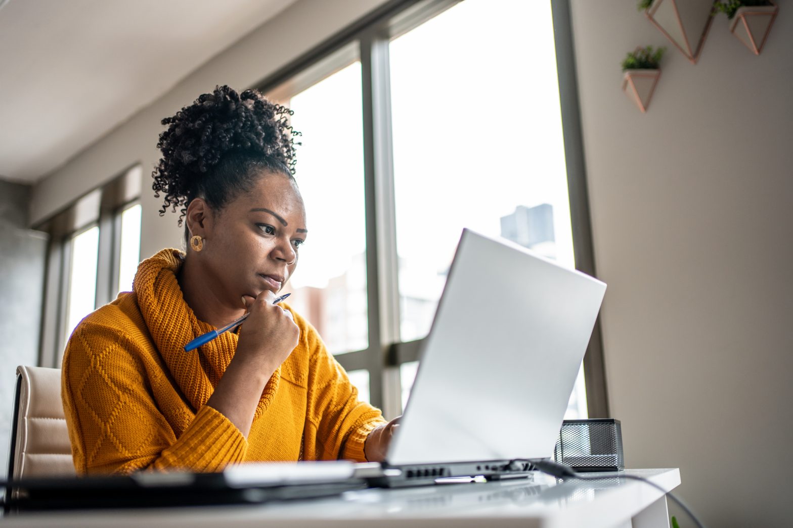Woman working from home office looking at a laptop with a pen in hand